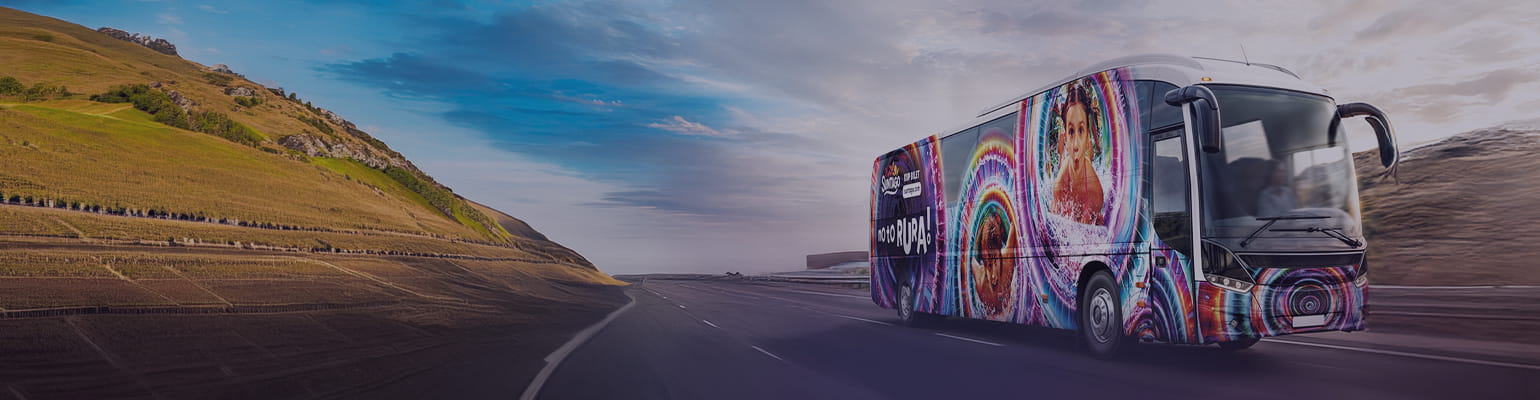 Suntago bus decorated with palm tree graphics. The side of the bus displays the text "Buy a ticket at suntago.com". In the background, silhouettes of palm trees against the setting sun.